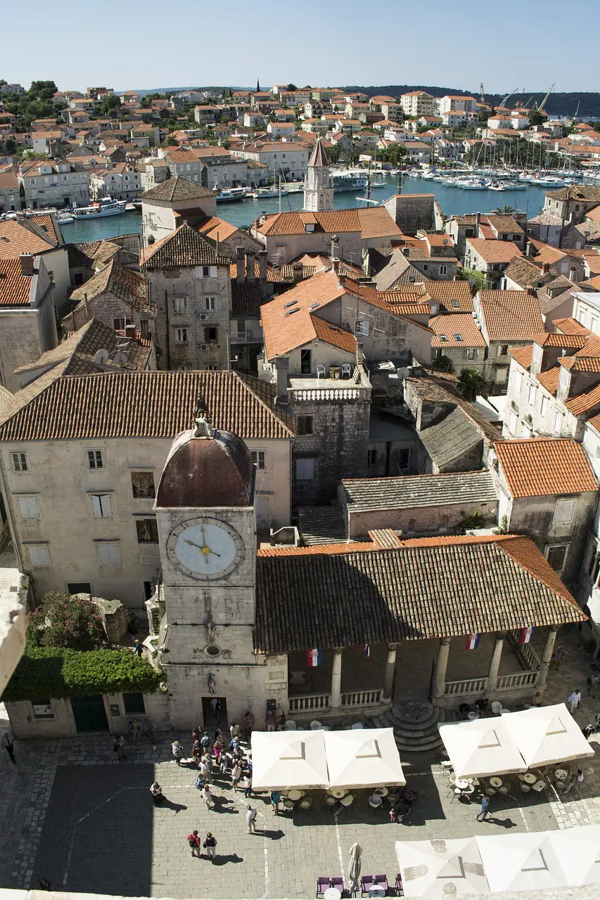 Aerial view of a historic European town with terracotta rooftops, a clock tower, and outdoor café seating; a river and distant marina are visible in the background—explore it all with Rewind Croatia tours.