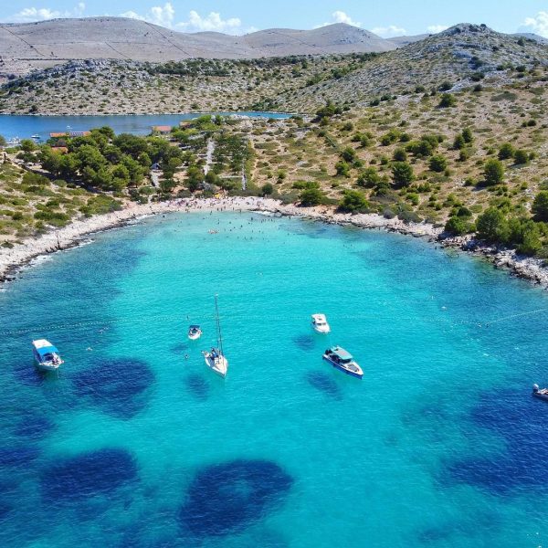Aerial view of a turquoise bay near Dubrovnik with several boats anchored and people swimming by a rocky shoreline with sparse vegetation, perfect for private boat tours or day trips.