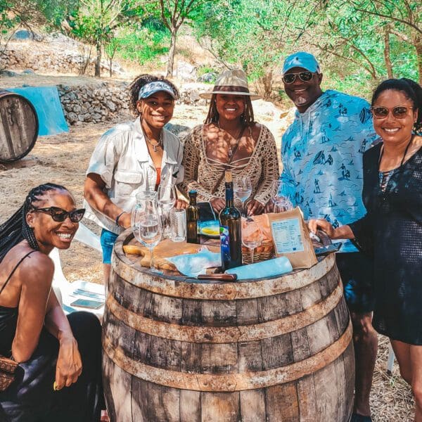 A group of people gather around a wooden barrel table outdoors, enjoying wine and snacks on a sunny day in Dubrovnik. Two wine barrels and greenery are seen in the background, reminiscent of delightful moments shared during private boat tours or day trips.