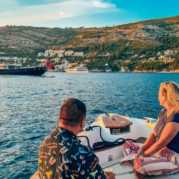 Two people sit on a small boat, observing a large yacht and other boats on the water, with hilly terrain and buildings in the background. It's one of those picturesque day trips you might take when visiting Dubrovnik, perhaps enjoying private boat tours to soak in all the breathtaking sights.