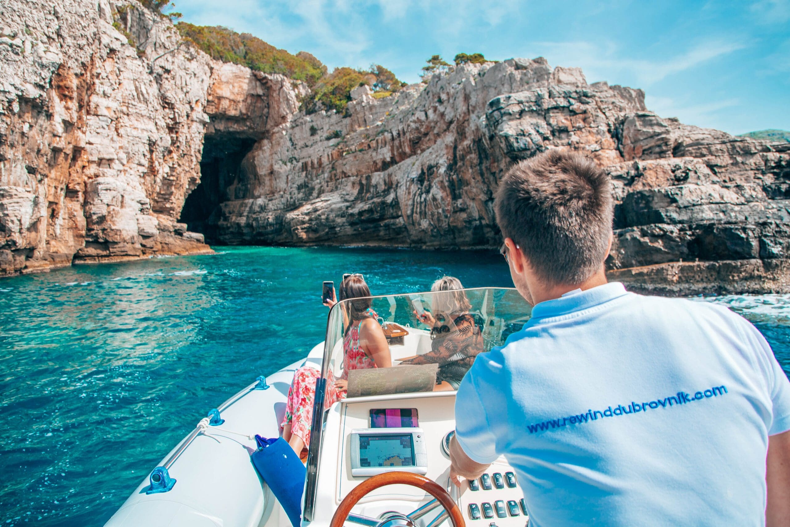 A person navigates a boat with two passengers toward a rocky cave along the turquoise coastline of Dubrovnik, making it an ideal setting for private boat tours.