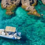 A small motorboat with a canopy floats on clear turquoise water near rocky cliffs in Dubrovnik, with a person swimming nearby, perfect for private boat tours and day trips.