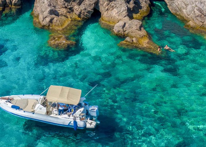 A small motorboat with a canopy floats on clear turquoise water near rocky cliffs in Dubrovnik, with a person swimming nearby, perfect for private boat tours and day trips.
