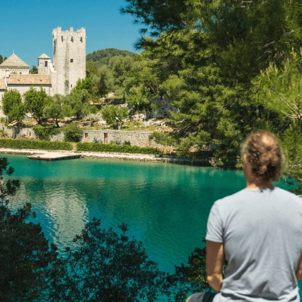 A person in a gray shirt is standing by a body of turquoise water, looking towards a historic stone building and tower surrounded by greenery, perhaps contemplating the charm of Dubrovnik's landscape on one of their private boat tours.