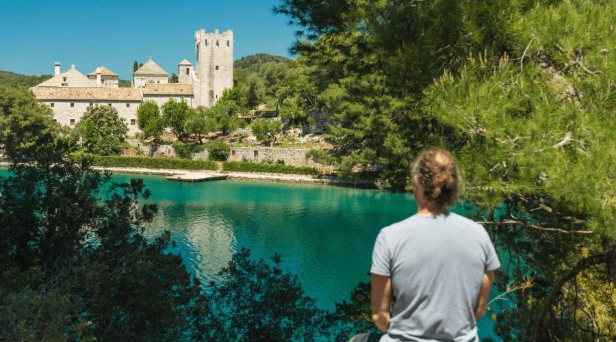 A person in a gray shirt is standing by a body of turquoise water, looking towards a historic stone building and tower surrounded by greenery, perhaps contemplating the charm of Dubrovnik's landscape on one of their private boat tours.