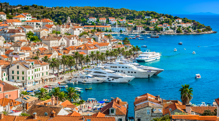 A seaside town with red-roofed buildings, palm trees, and several yachts docked in a vibrant blue bay under a clear sky. Hills and more buildings are in the background, perfect for day trips or private boat tours to explore the stunning surroundings of Dubrovnik.