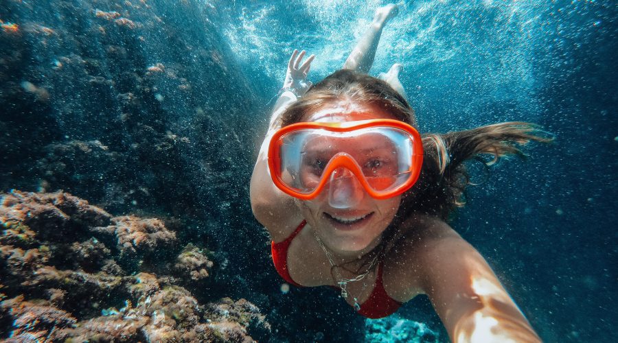 A person wearing red swim goggles and a matching red swimsuit is underwater, swimming near a coral reef off the coast of Dubrovnik. They are smiling and taking a selfie while enjoying one of their unforgettable private boat tours.