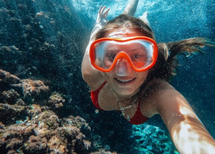 A person wearing red swim goggles and a matching red swimsuit is underwater, swimming near a coral reef off the coast of Dubrovnik. They are smiling and taking a selfie while enjoying one of their unforgettable private boat tours.