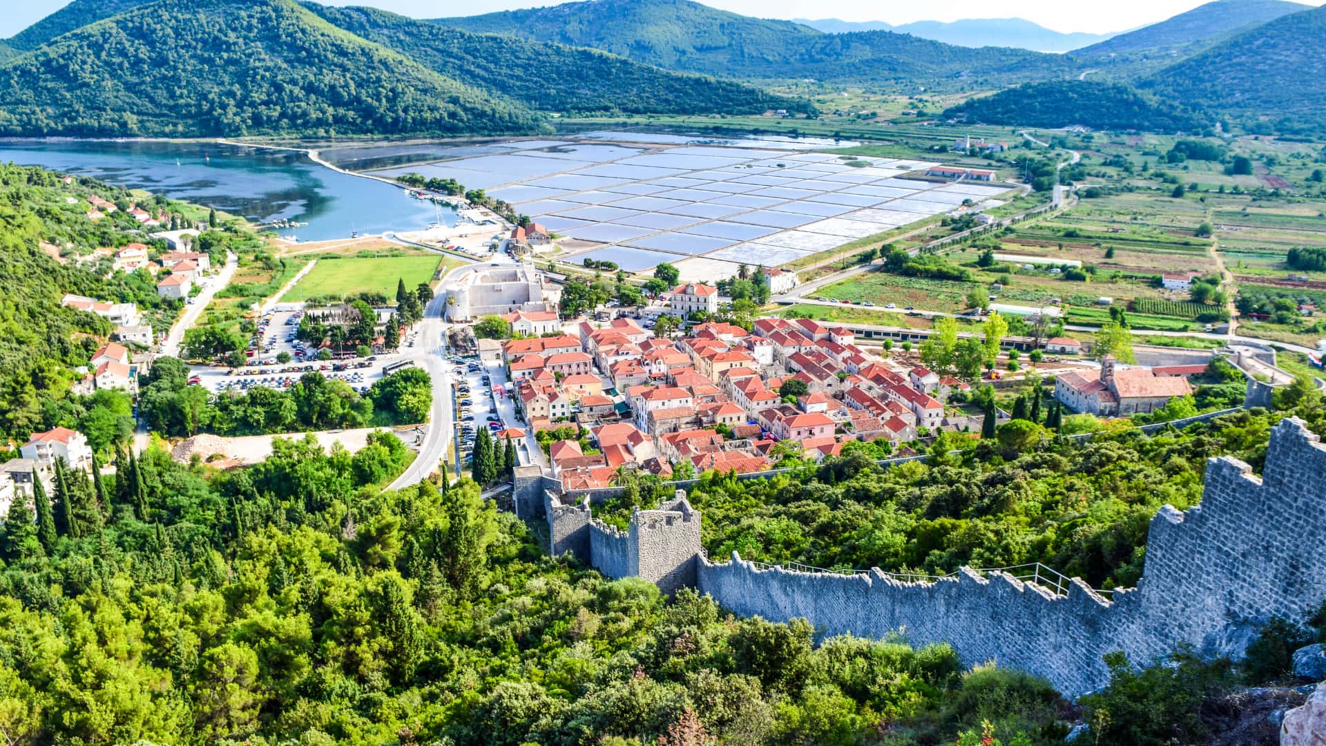 Aerial view of the medieval town Ston, surrounded by lush greenery and mountains. The scene captures orange-roofed buildings, a stone wall, and a rectangular grid of salt pans in the background, inviting exploration by boat for a unique perspective.