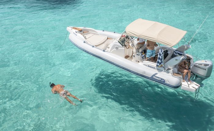 A person swims near a small motorboat on clear turquoise water, part of one of Dubrovnik's private boat tours. Several others stay on the boat, with one using binoculars. The vessel is equipped with a Honda outboard motor and a sunshade.