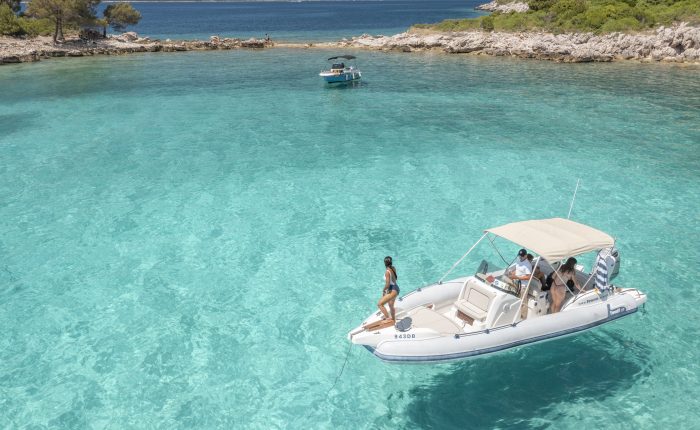A white motorboat with a canopy floats on clear turquoise water near a rocky shore with greenery, offering an idyllic setting for private boat tours in Dubrovnik. Another boat can be seen in the background, and people are relaxing on the first boat, enjoying their day trip.