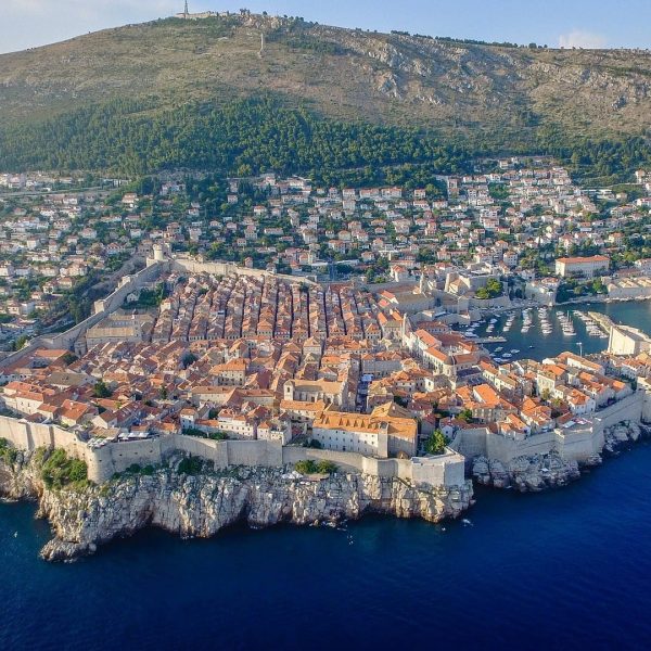 Aerial view of Dubrovnik, a coastal town with an old walled city featuring orange-roofed buildings, surrounded by rocky cliffs and water, set against a hilly backdrop. Perfect for day trips and private boat tours.