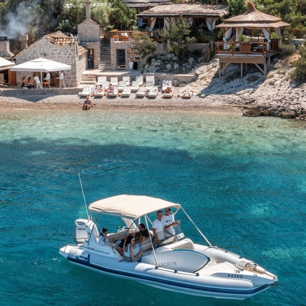 A small motorboat with people on board is on clear blue waters near a rocky shoreline, perfect for private boat tours. Several buildings and a restaurant with outdoor seating are visible in the background, making it an ideal spot for day trips in Dubrovnik.