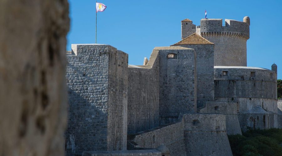 A view of historic stone fortress walls with a turret, a flag flying on top, under a clear blue sky, captures the essence of Dubrovnik. Perfect for day trips or private boat tours exploring this majestic coastal city.
