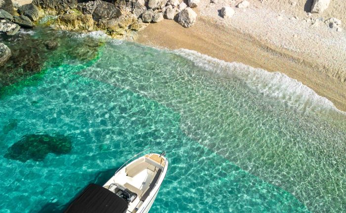 A boat with a black canopy is anchored in clear turquoise waters near a rocky shoreline with a sandy beach, perfect for day trips or private boat tours around Dubrovnik.