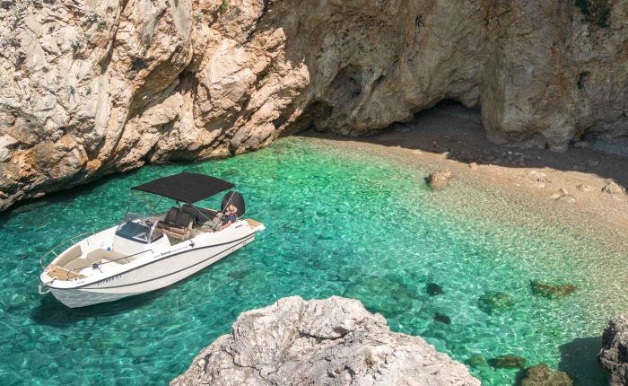 A small white boat with a black canopy is anchored in clear turquoise water near a rocky cliff and a small beach with large rocks, perfect for Private Boat Tours in Dubrovnik.