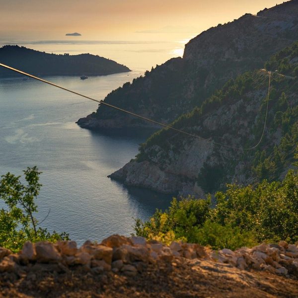 A coastal landscape at sunset near Dubrovnik, featuring hills with lush greenery, a winding road perfect for day trips, and islands in the distance. Cable lines run through the tranquil scene above calm waters, ideal for private boat tours.