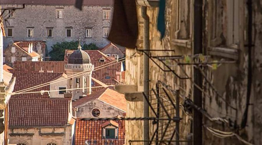 Clothes hanging on a line over a narrow, historic street with old buildings and a dome in the background, likely in Dubrovnik—a picturesque Mediterranean city known for its private boat tours.