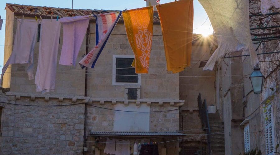 Clothes are hanging on lines strung between buildings in a sunlit courtyard in Dubrovnik. A flag is visible on the roof of one of the buildings, adding to the charm of this historic city that's perfect for private boat tours and day trips.