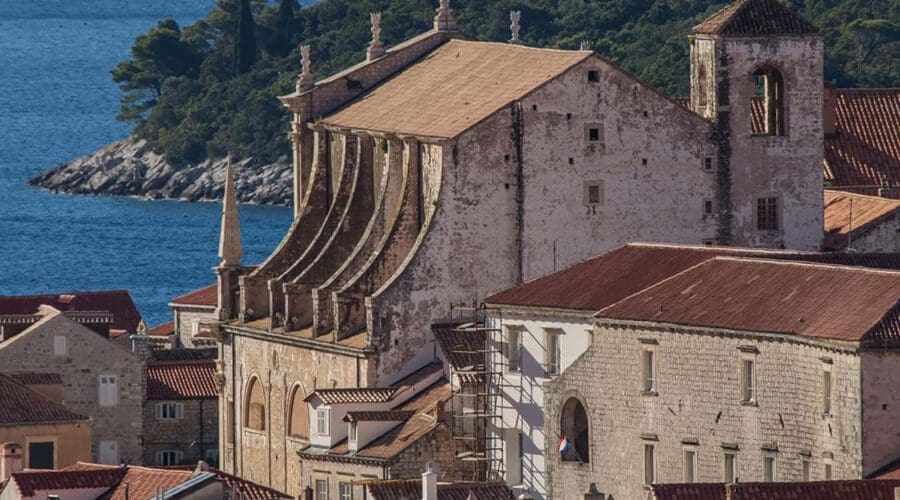 Historic seaside town with red-tiled roofs, a large stone church, and the Adriatic Sea in the background with lush green hills; Dubrovnik offers private boat tours and day trips for those seeking to explore its scenic beauty.