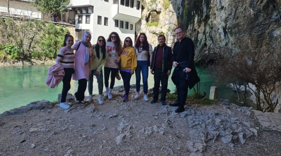 A group of eight people stand on a rocky ledge by a scenic river in Dubrovnik with a traditional white building and cliffside in the background, perfect for memorable day trips.