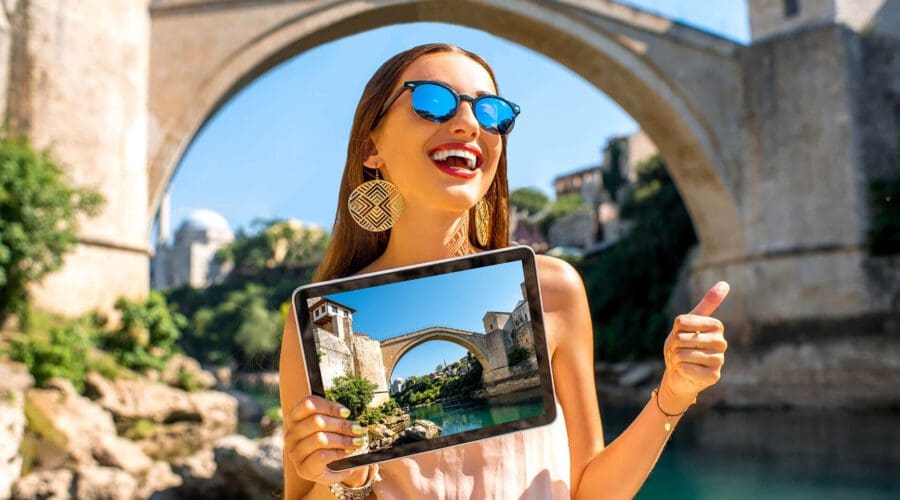 Woman holding a tablet displaying an image of a bridge in Dubrovnik while posing in front of the same bridge. She is wearing sunglasses and giving a thumbs up, showcasing the perfect spot for unforgettable day trips.
