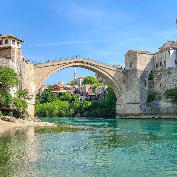 A stone arch bridge spans a river between historical buildings on both sides, under a clear blue sky with green water and lush vegetation along the riverbanks, much like the picturesque scenes you would encounter on Dubrovnik day trips or private boat tours.