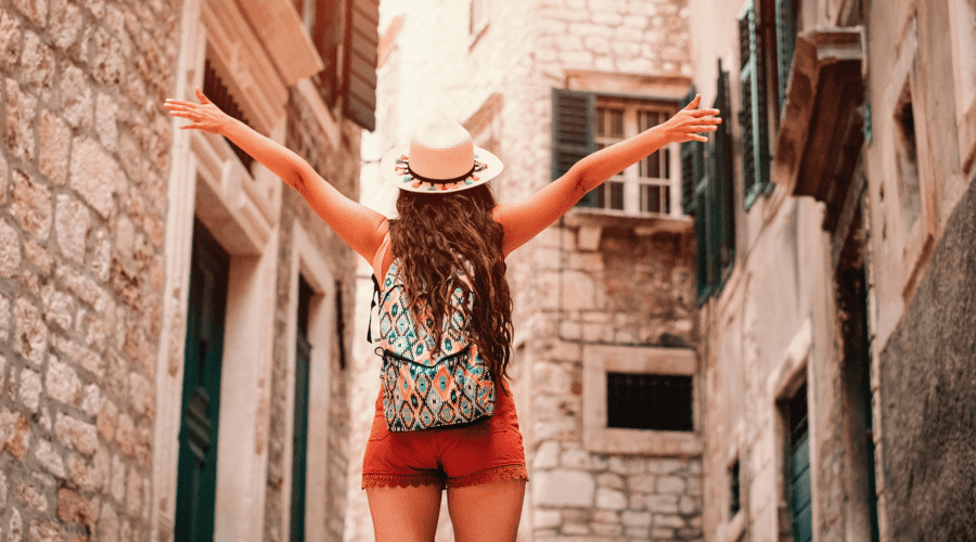 A person wearing a hat and backpack stands with arms outstretched in a narrow alley between stone buildings, perhaps reveling in the charm of Dubrovnik before embarking on one of many day trips or private boat tours.