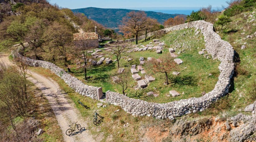 An aerial view of an ancient stone cemetery enclosed by a wall, with a dirt path and two bicycles parked outside. Trees and a mountain landscape are visible in the background, perfect for those exploring Dubrovnik on day trips or private boat tours.