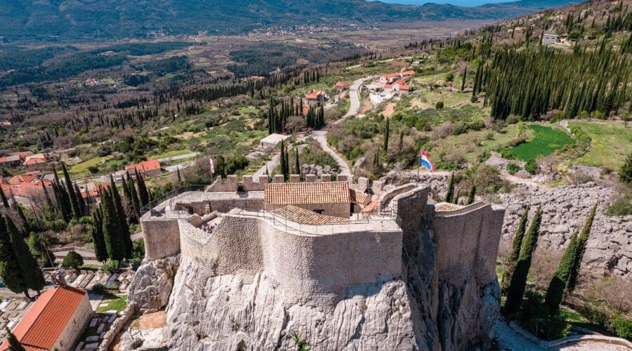 Aerial view of an old stone fortress perched on a rocky hill, reminiscent of Dubrovnik, surrounded by a countryside landscape with scattered buildings, trees, and winding roads—an idyllic scene perfect for day trips or private boat tours.