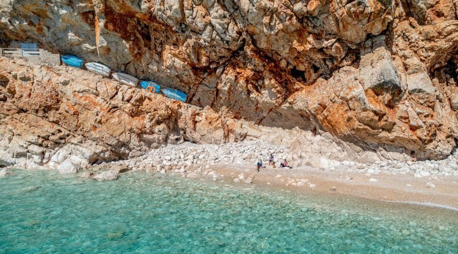 A rocky cliffside with multiple boats stored on racks above a small, isolated beach. Clear turquoise water laps against the shore in Dubrovnik, where a few people are gathered on the sand and rocks—perfect for private boat tours and day trips.