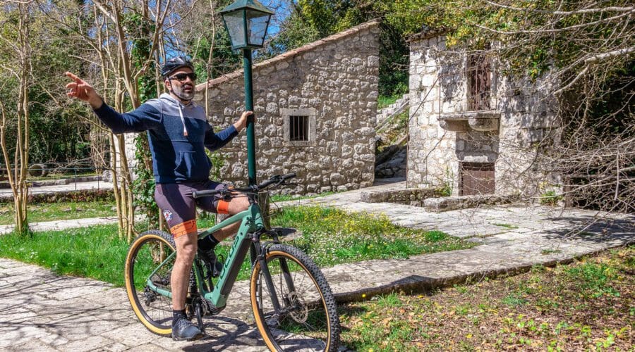 A man stands astride a bicycle, pointing to the side. He is wearing a hoodie, shorts, and sunglasses. Behind him are stone buildings and greenery under a sunny sky, typical of Dubrovnik's charm—a perfect spot for day trips or private boat tours.