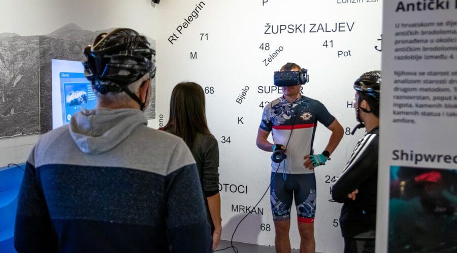 A group of people, some wearing bicycle helmets, observes a man using a VR headset in a modern exhibit room with maps and information about Dubrovnik's private boat tours and day trips displayed on the walls.