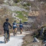 Three cyclists ride on a gravel path through a rocky landscape with sparse vegetation. An old stone building with a tiled roof is visible in the background, reminiscent of the charming outskirts of Dubrovnik that are perfect for day trips.