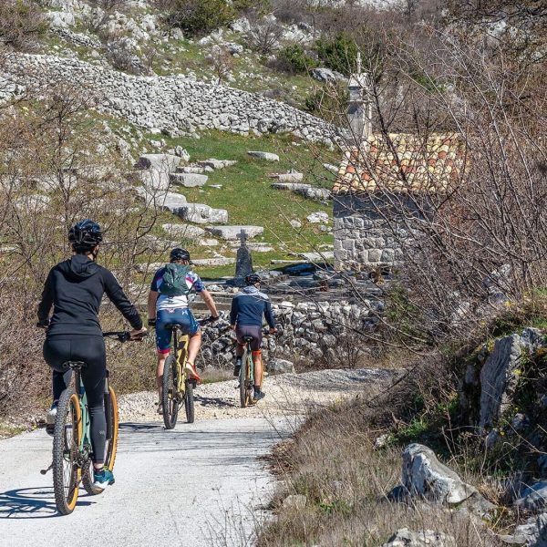 Three cyclists ride on a gravel path through a rocky landscape with sparse vegetation. An old stone building with a tiled roof is visible in the background, reminiscent of the charming outskirts of Dubrovnik that are perfect for day trips.