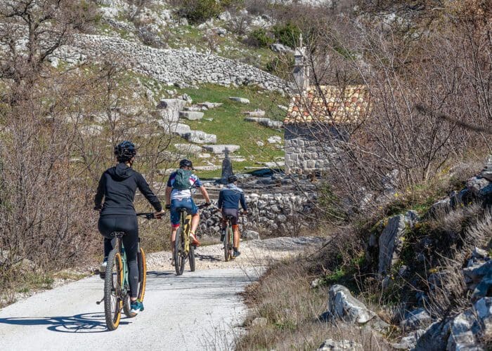 Three cyclists ride on a gravel path through a rocky landscape with sparse vegetation. An old stone building with a tiled roof is visible in the background, reminiscent of the charming outskirts of Dubrovnik that are perfect for day trips.