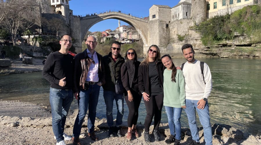 A group of seven people stands by a riverside with a historic stone bridge and buildings in the background, posing for a photo in casual attire on a sunny day, enjoying their day trip to Dubrovnik.