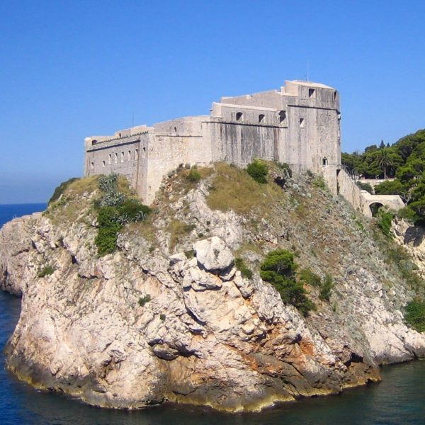 A large stone fortress sits atop a rocky cliff, overlooking the blue sea near Dubrovnik, surrounded by some greenery under a clear blue sky – an ideal spot for day trips or private boat tours.