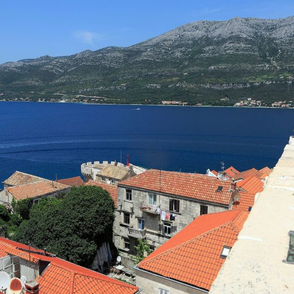 A scenic view of Dubrovnik with its iconic orange-tiled rooftops, blue sea, and distant mountains under a clear sky. Ideal for day trips or private boat tours, this coastal town offers breathtaking beauty at every turn.