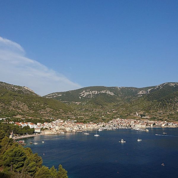 A coastal town with red-roofed buildings is nestled between green hills and a calm blue sea, perfect for day trips near Dubrovnik. Several boats are anchored in the water, hinting at the potential for private boat tours on a clear, sunny day.
