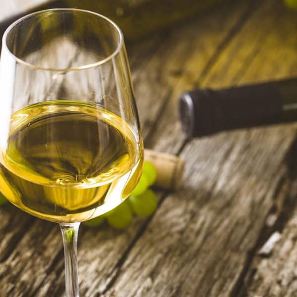 A glass of white wine on a wooden table with wine bottles, corks, and green grapes in the background evokes memories of private boat tours around Dubrovnik.