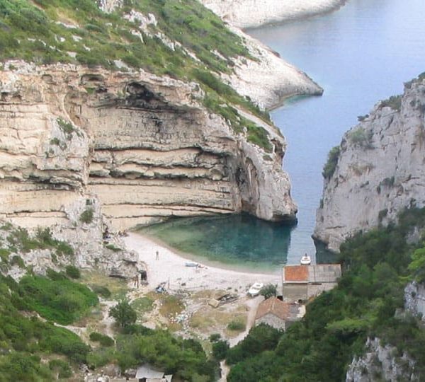A secluded, narrow cove with clear blue water surrounded by high, rocky cliffs and lush greenery awaits you on one of our private boat tours from Dubrovnik. A small building with a red roof sits quaintly near the water's edge, perfect for those idyllic day trips.