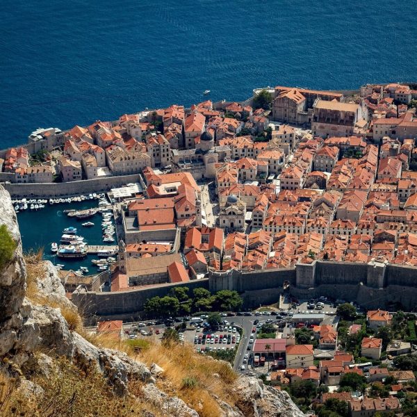 Aerial view of Dubrovnik, a coastal town with numerous orange-roofed buildings, a marina with boats, and a stone wall surrounding the area, set against a backdrop of the sea. Perfect for day trips and private boat tours.