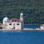 A stone building with a dome and bell tower sits on a small island surrounded by the blue waters of the Adriatic, with a forested hillside in the background, capturing the serene beauty reminiscent of Croatia's stunning landscapes.