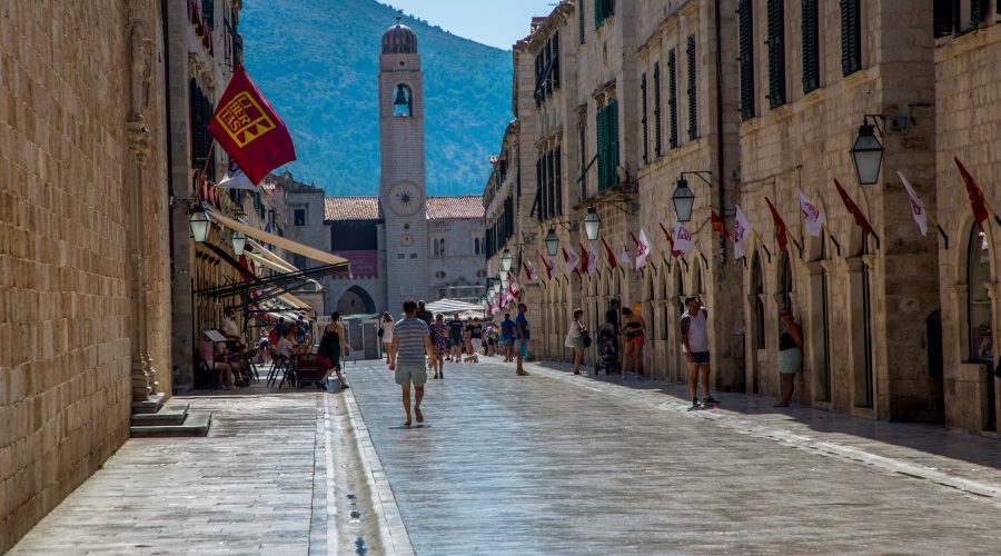 A narrow, sunlit stone street lined with buildings and flags guides adventurers toward a clock tower under a clear blue sky. Experience the Pearl of the Adriatic as a few people leisurely stroll along this iconic path in Dubrovnik.
