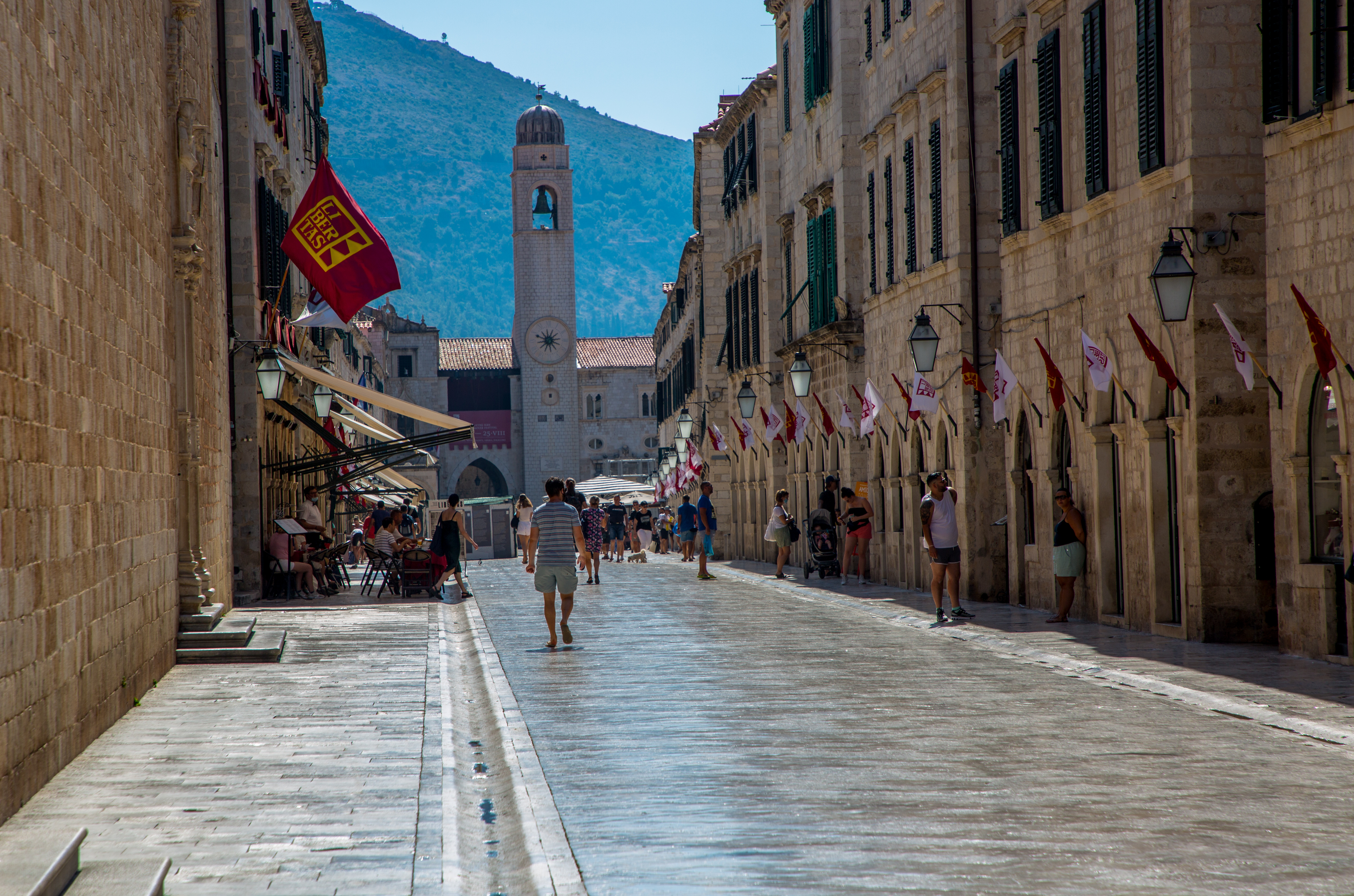 A narrow, sunlit stone street lined with buildings and flags guides adventurers toward a clock tower under a clear blue sky. Experience the Pearl of the Adriatic as a few people leisurely stroll along this iconic path in Dubrovnik.