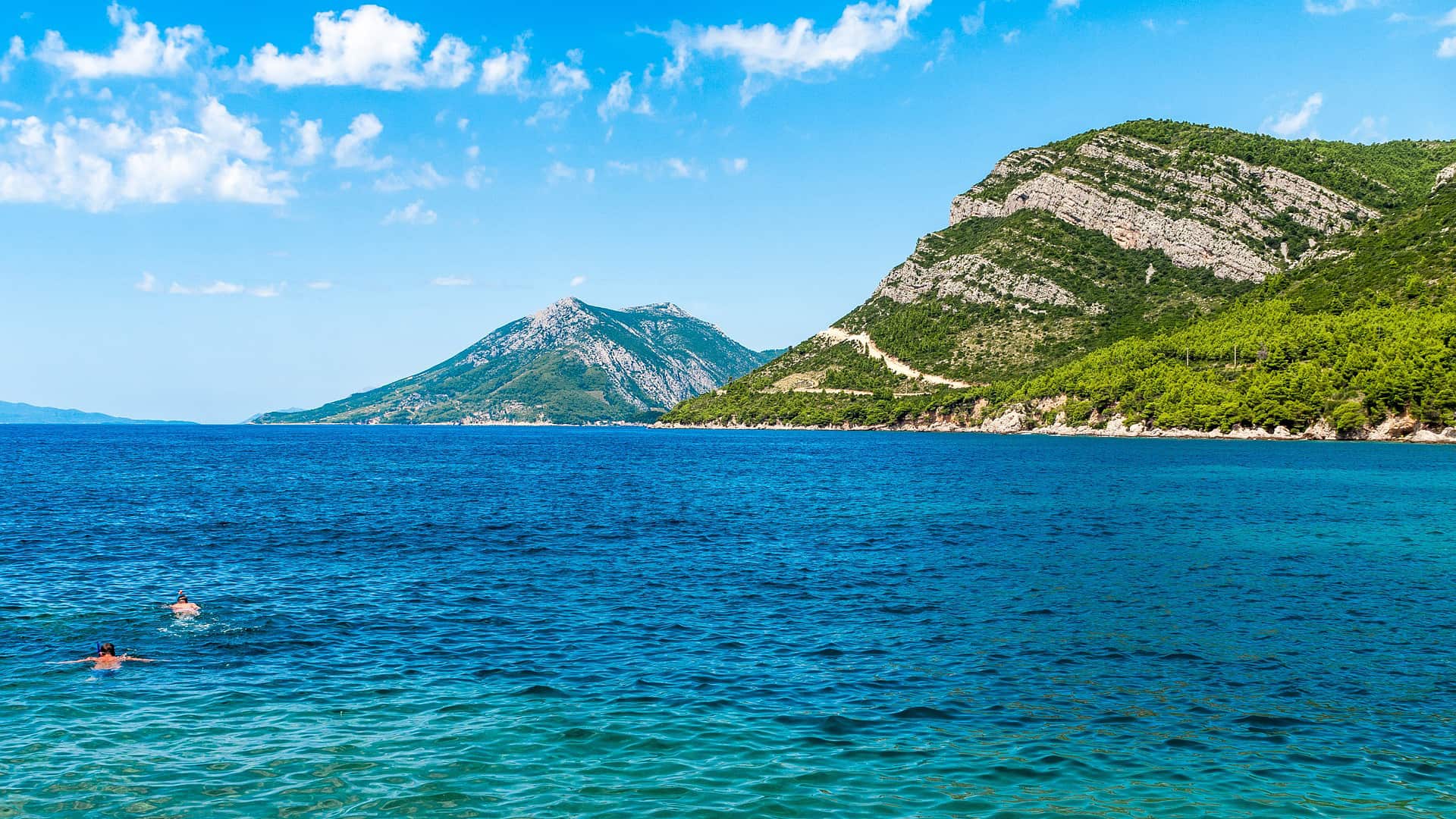 Two people swim in the clear blue waters near a green mountainous shoreline under a bright blue sky with scattered clouds, enjoying one of Dubrovnik's private boat tours.