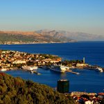 A coastal city with a harbor and several docked cruise ships, surrounded by mountainous terrain and greenery under a clear blue sky, Dubrovnik offers scenic day trips and exclusive private boat tours.