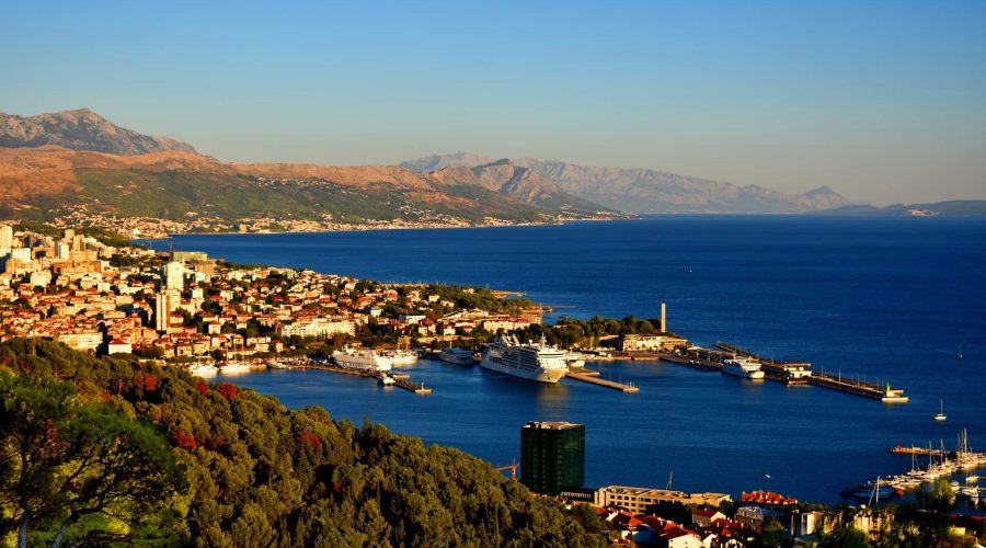 A coastal city with a harbor and several docked cruise ships, surrounded by mountainous terrain and greenery under a clear blue sky, Dubrovnik offers scenic day trips and exclusive private boat tours.