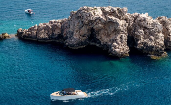 Two boats are on clear blue water near a rocky outcrop in Dubrovnik. A smaller boat, part of the private boat tours, is docked close to the rocks, while a larger motorboat cruises past.